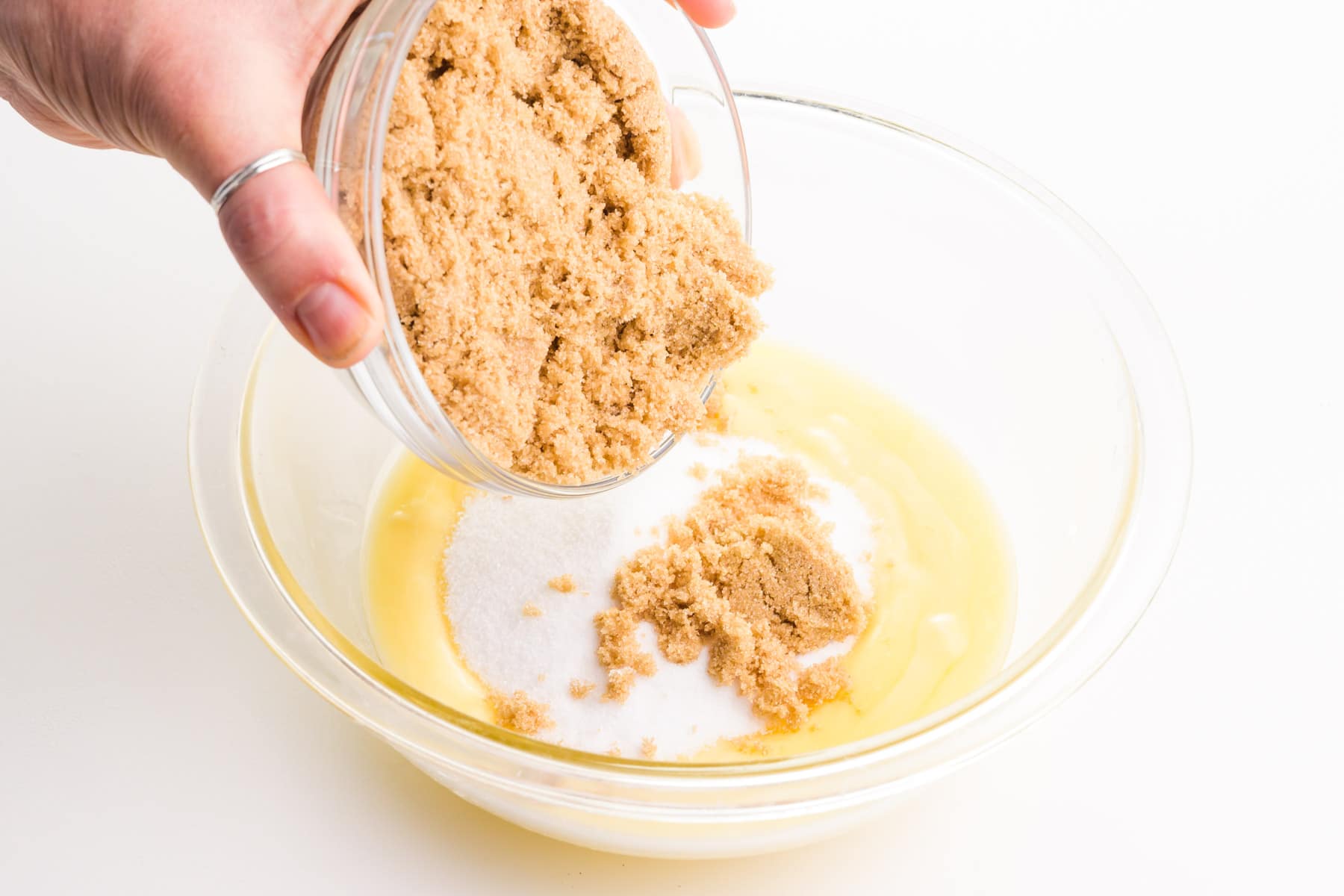 A hand holds a bowl of brown sugar, pouring it into a bowl with melted butter and granulated sugar.