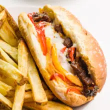 A closeup of a vegan Philly cheesesteak sandwich sitting next to French fries on a plate.