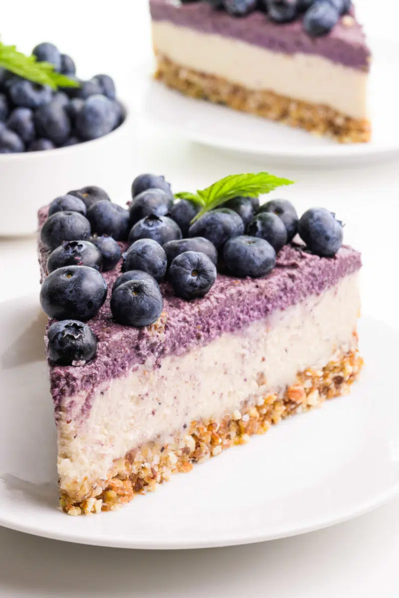A slice of raw cheesecake has blueberries on top. It's sitting on a white plate. Another slice and a bowl of berries is in the background.