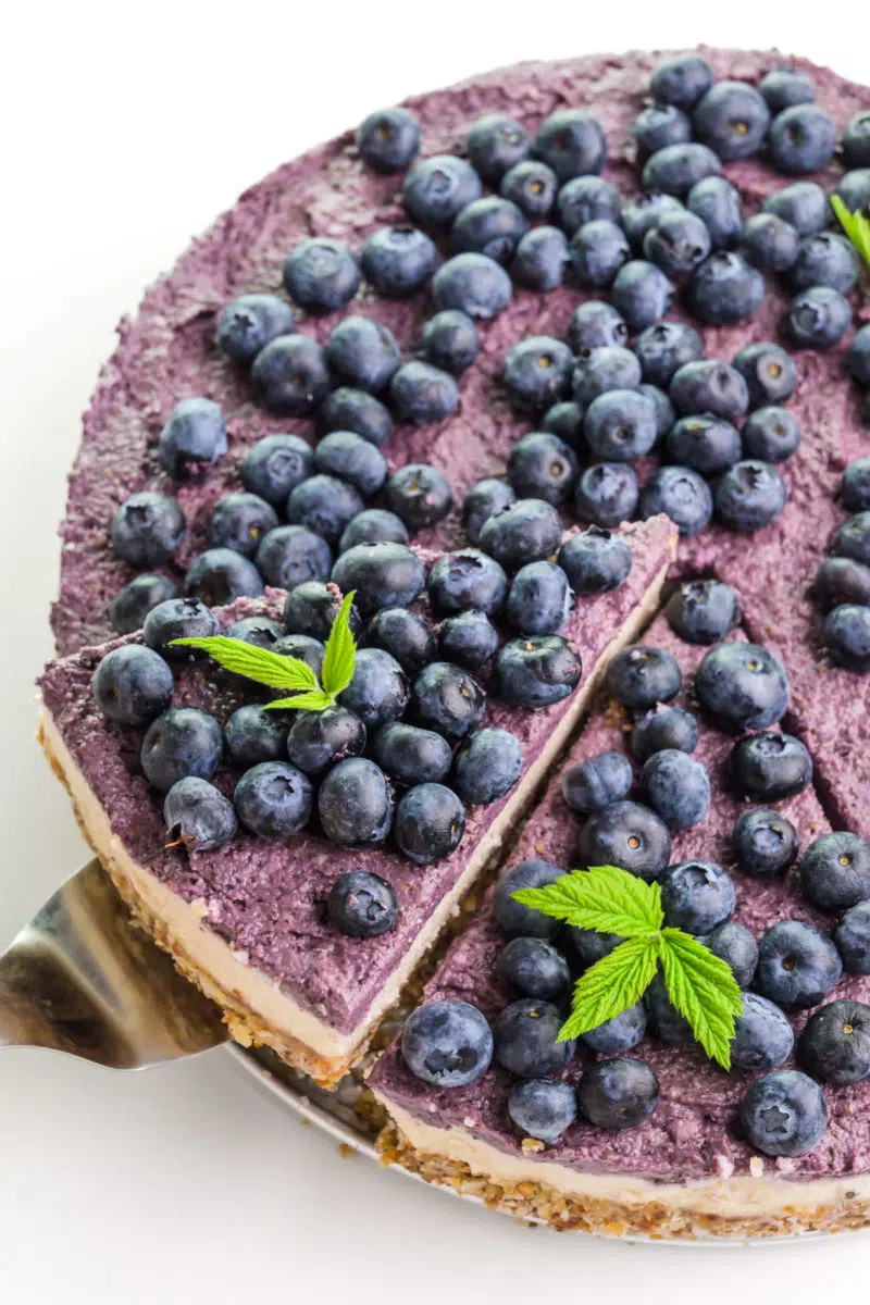 Looking down on a vegan raw cheesecake with fresh blueberries on top. A slice is being lifted out with a spatula.