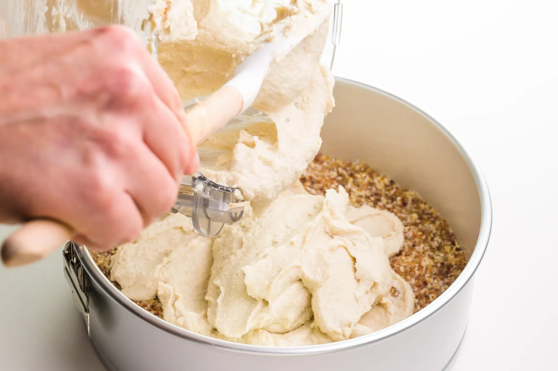 A hand uses a spatula to spoon cashew filling from a food processor to a springform pan.