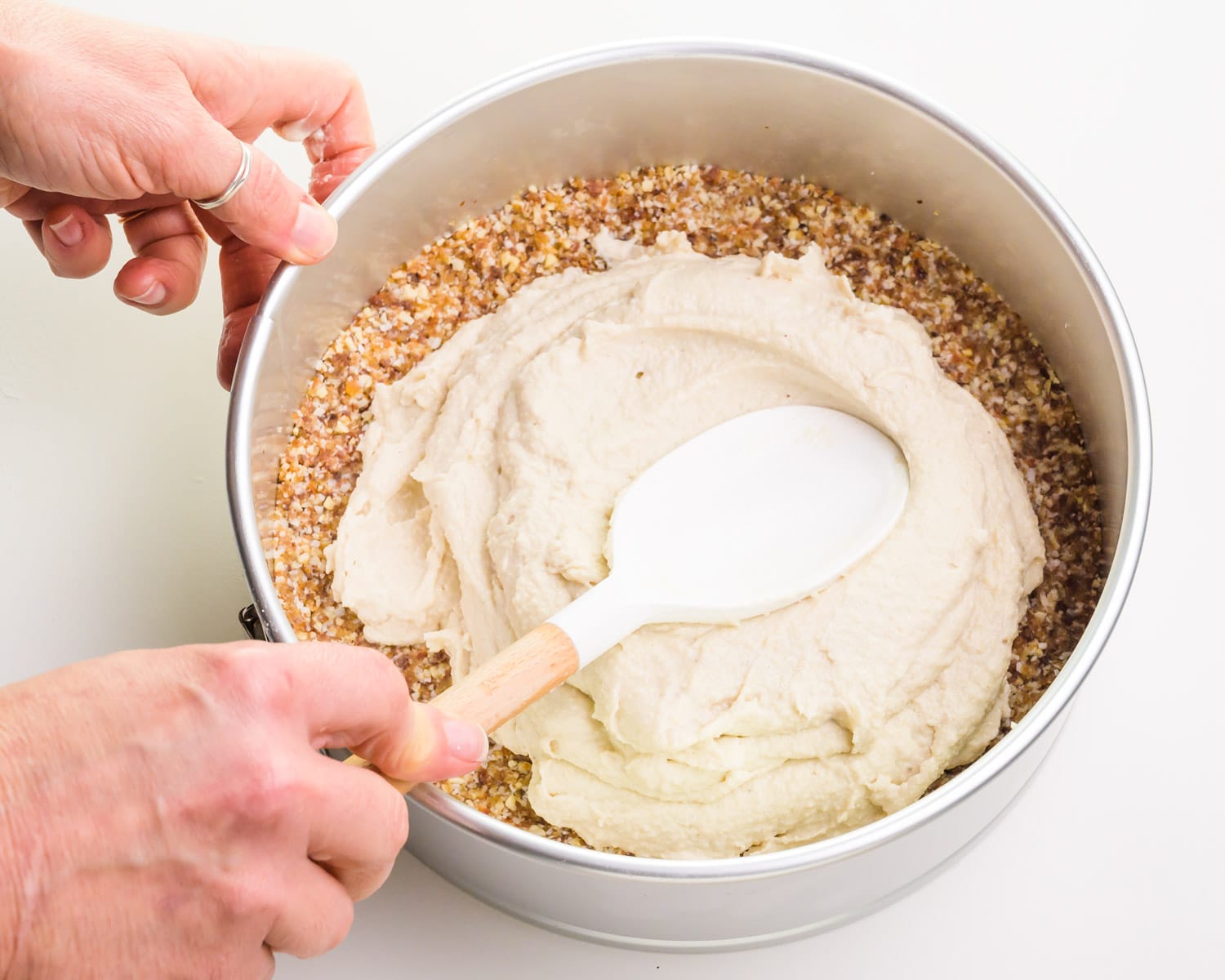 A hand holds a spatula, spreading raw cashew filling evenly across the bottom of a springform pan.