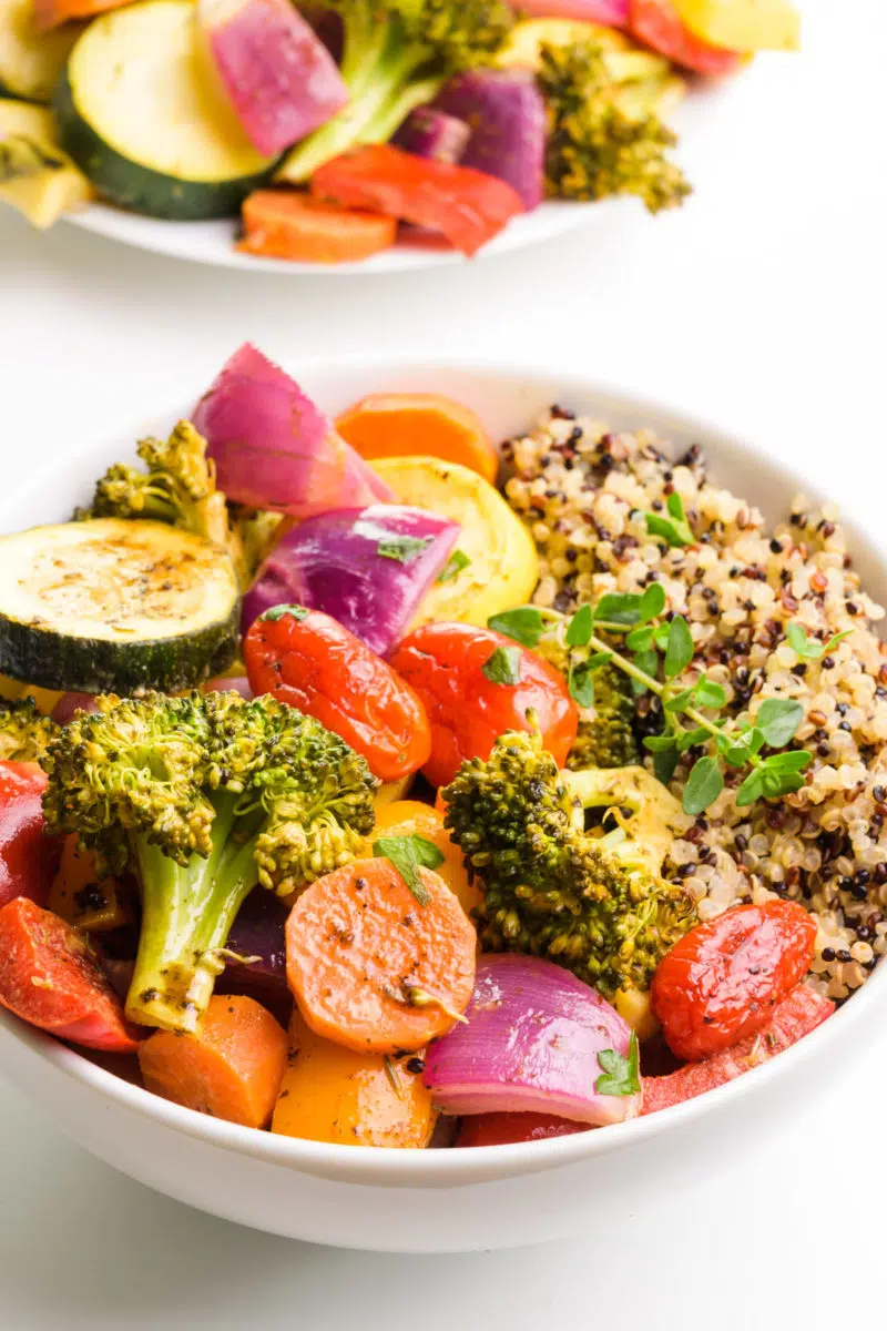 A bowl of vegan roasted veggies has quinoa on the side. There is a plate of vegetables in the background.