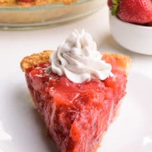 A slice of vegan strawberry pie sits on a plate. It has whipped cream on top. There is more pie and a bowl of strawberries in the background.