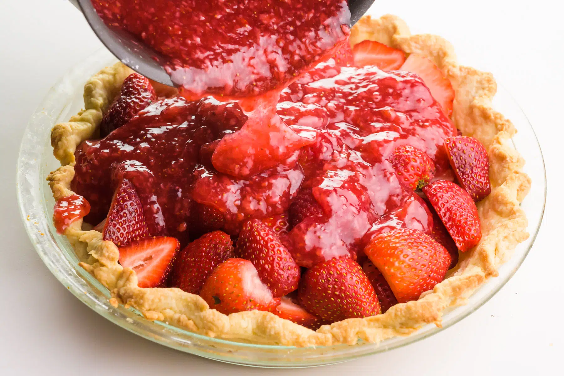 A thick strawberry gel is being poured over fresh strawberries in baked pie crust.