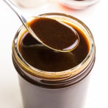 A spon hovers over a mason jar full of vegan Worcestershire sauce. A few cloves of garlic and a bowl of soy sauce sits in the background.