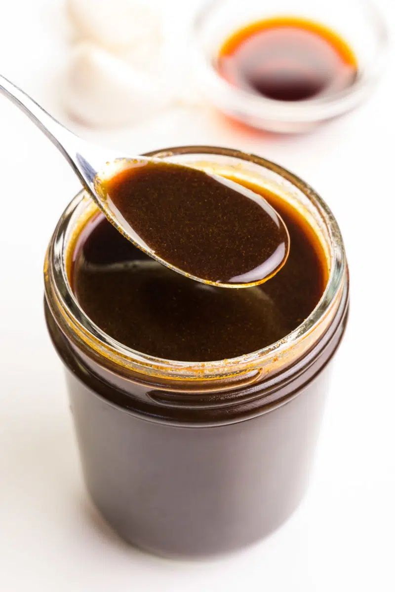A spon hovers over a mason jar full of vegan Worcestershire sauce. A few cloves of garlic and a bowl of soy sauce sits in the background.