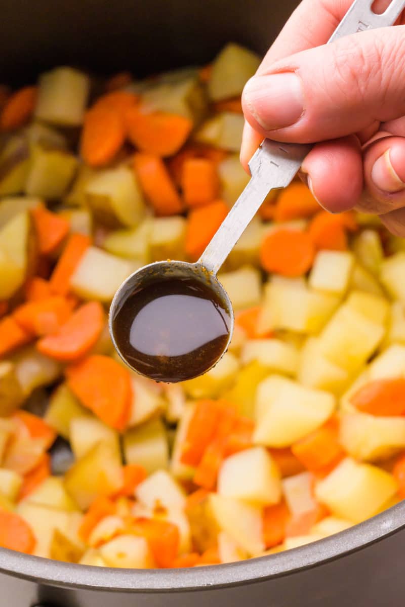 A hand holds a spoonful of Worcestershire sauce over a pot of chopped veggies. 
