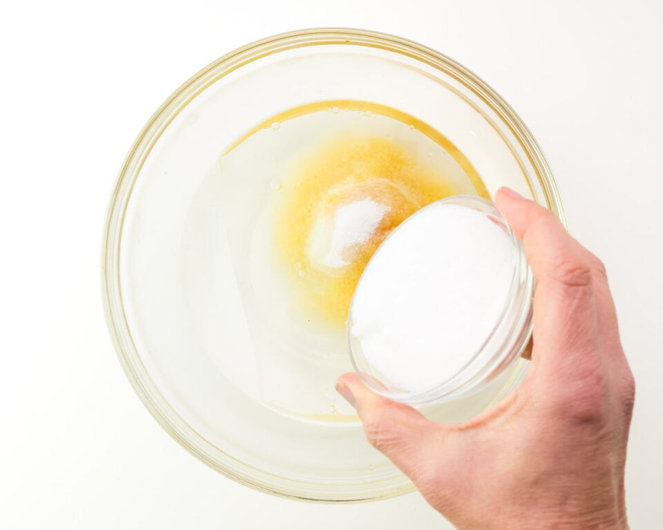 A hand holds a jar of white mixture, pouring it into a bowl with other liquid ingredients.
