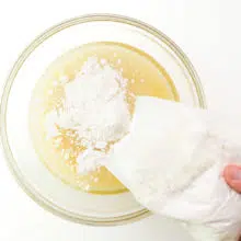 A hand holds a plastic bag of cake mix, pouring it into a mixing bowl with other ingredients.