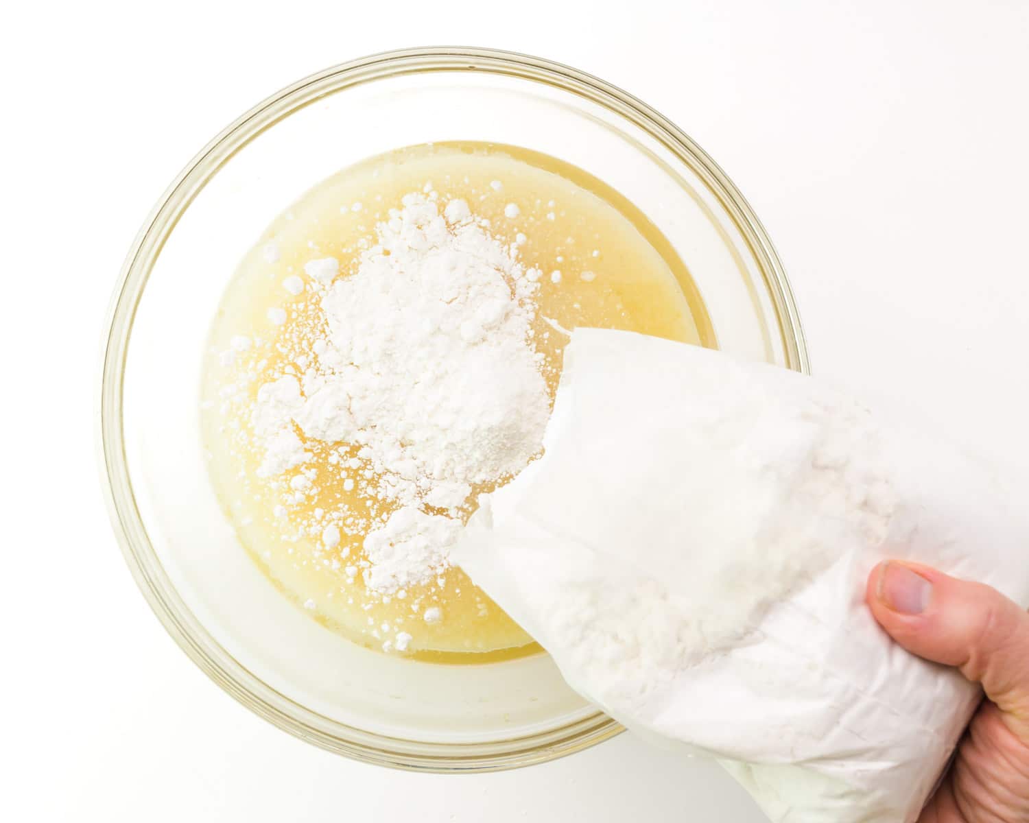 A hand holds a plastic bag of cake mix, pouring it into a mixing bowl with other ingredients.