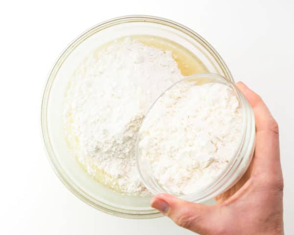 A hand pours a bowl of flour into a larger bowl with other ingredients.