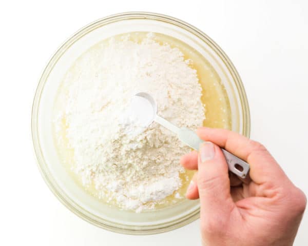 A hand holds a measuring spoon of baking powder, pouring it into a mixing bowl with other ingredients.