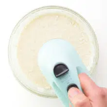 A hand holds a mixer, using it to mix cake batter in a mixing bowl.