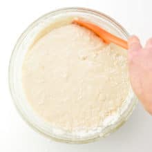 A hand holds an orange spatula, using it to scrape down the sides of a mixing bowl full of cake batter.