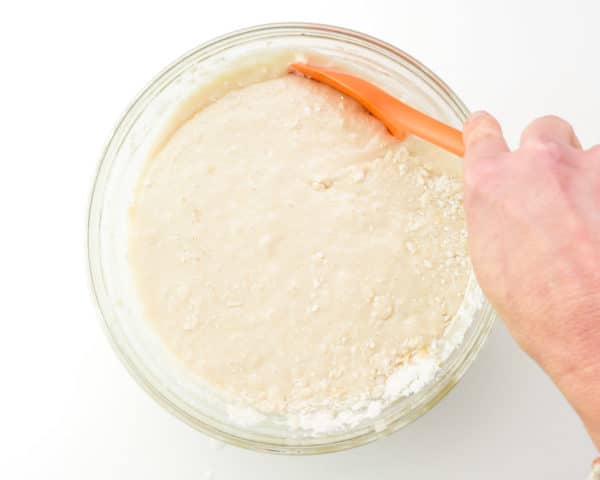 A hand holds an orange spatula, using it to scrape down the sides of a mixing bowl full of cake batter.