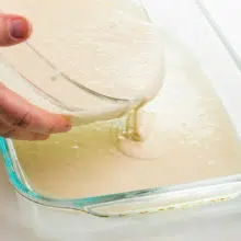 A hand holds a bowl, pouring cake batter into a glass baking dish.