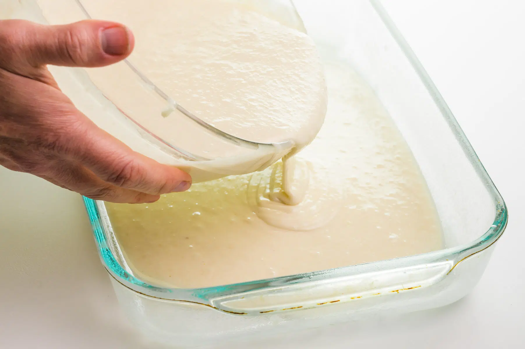 A hand holds a bowl, pouring cake batter into a glass baking dish.