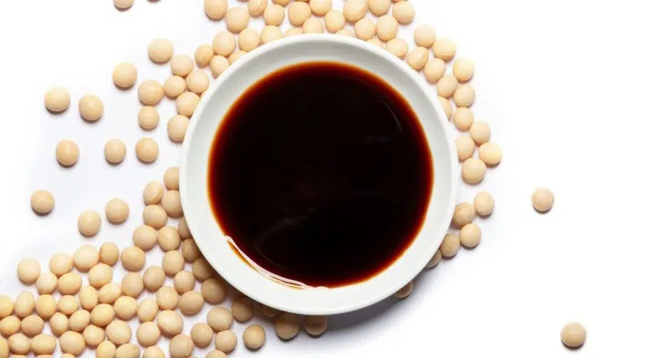 A bowl of tamari sauce sits on a white table, surrounded by soybeans.