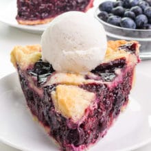 A slice of vegan berry pie sits on a plate. It has ice cream on top. There is another slice behind it and a bowl of fresh blueberries.