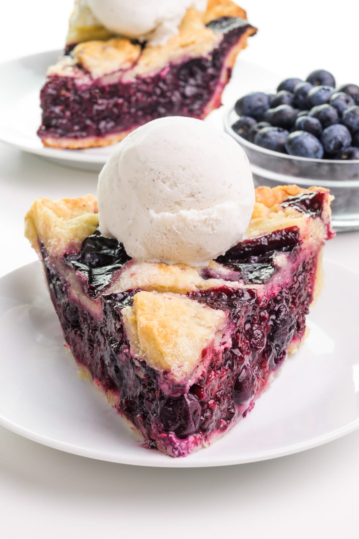 A slice of vegan berry pie sits on a plate.  There is ice cream on top.  Behind it is another slice and a bowl of fresh blueberries.