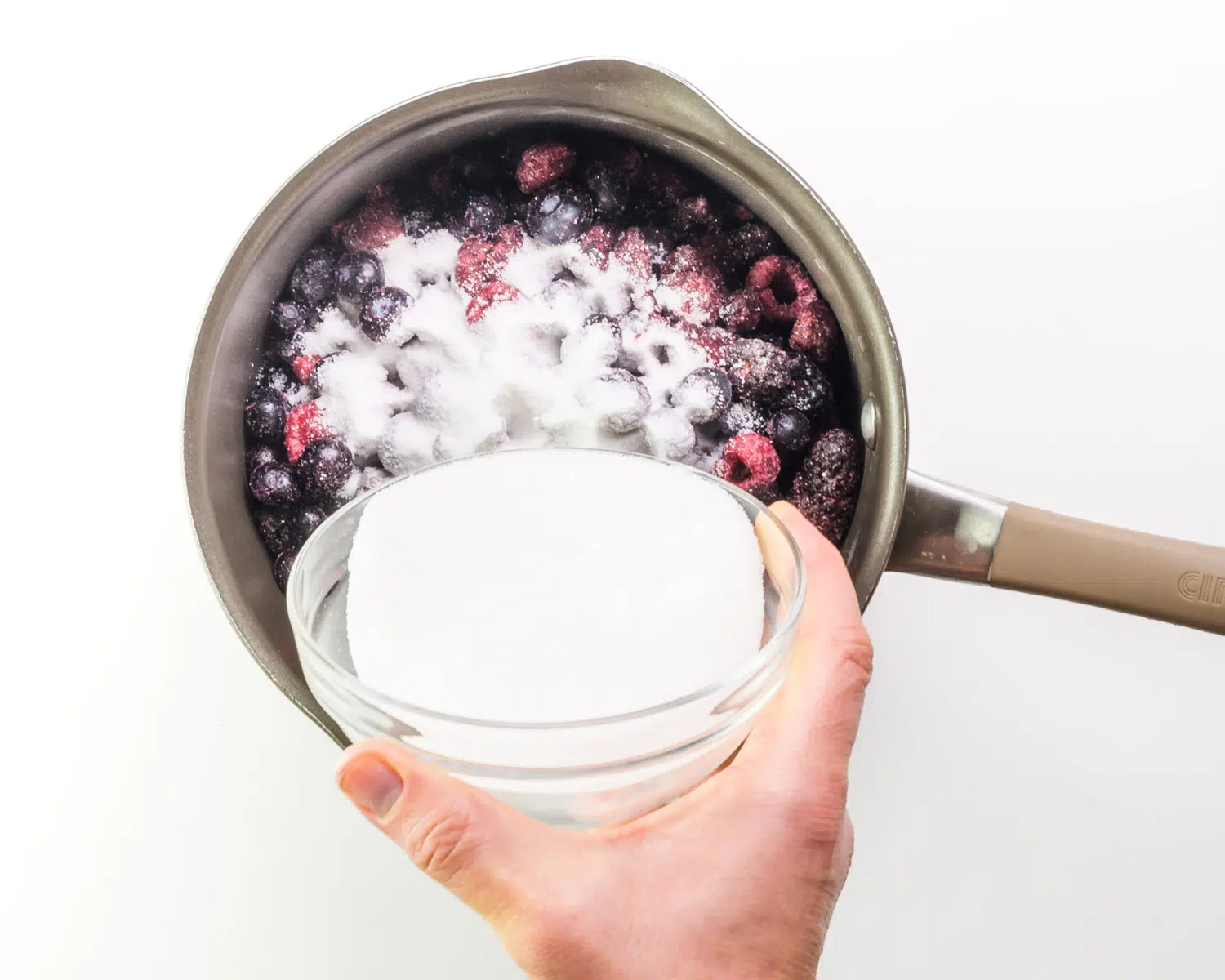 Sugar is being poured into a saucepan with mixed berries.