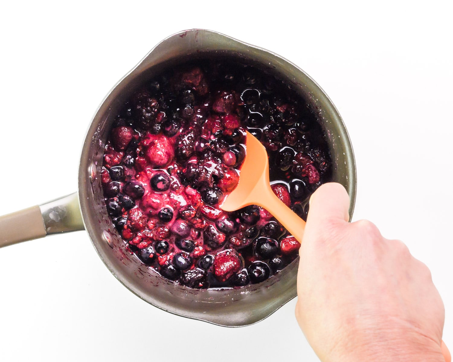 A hand holds a spatula, using it to stir berries in a saucepan.