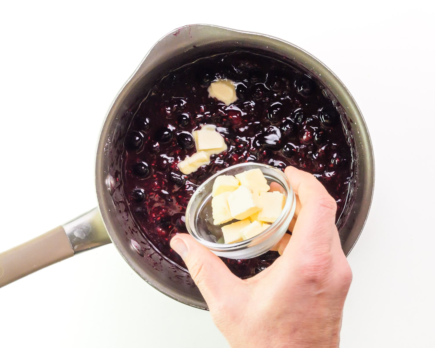 Vegan butter being poured from a bowl into a saucepan with berry sauce.