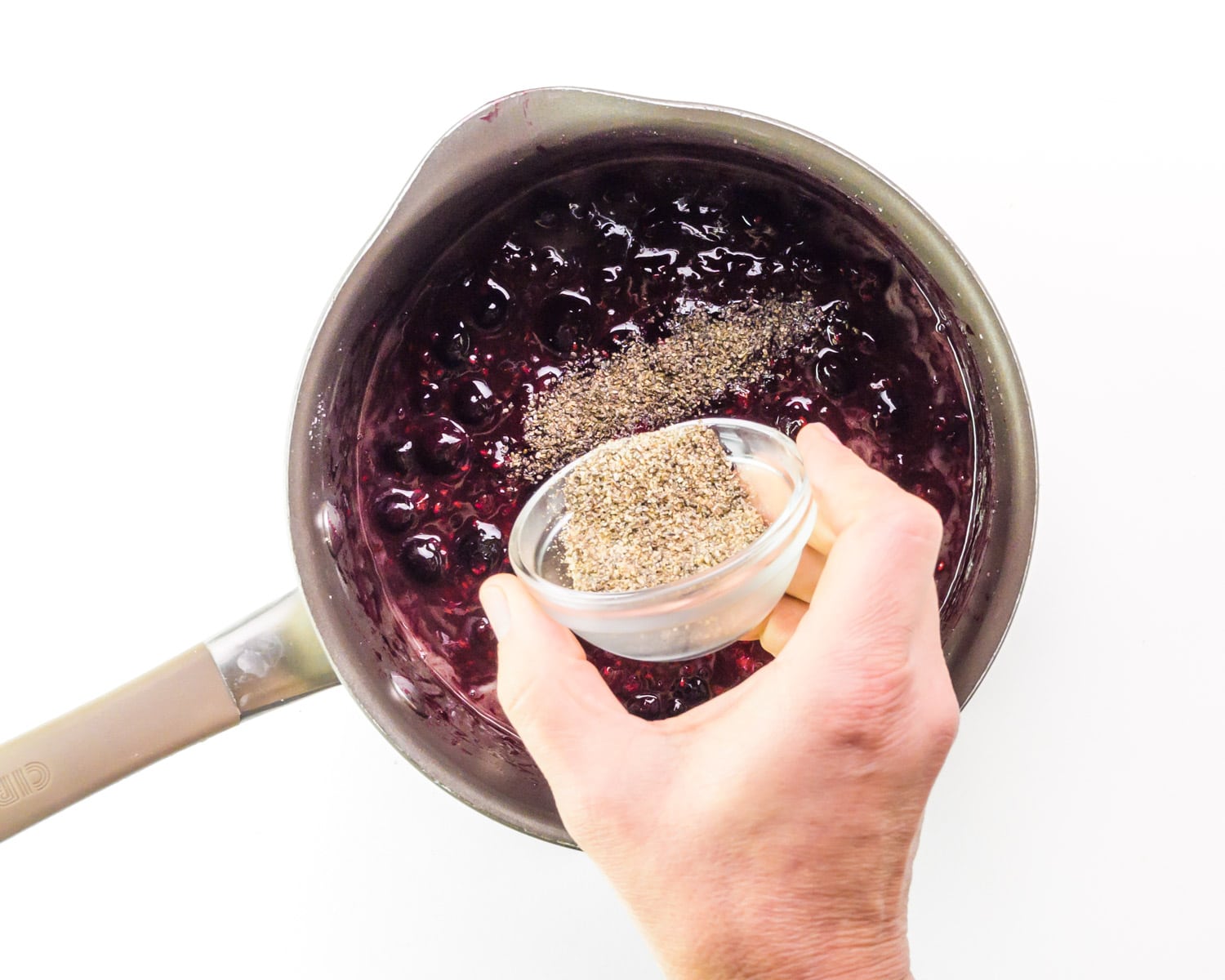Ground chia seeds being poured into a saucepan with a berry sauce.