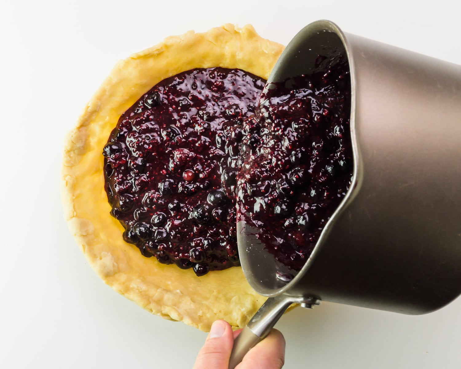 Berry filling is being poured from a saucepan into a prepared pie crust.