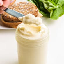 A hand holds a scoop of mayo on a knife, hovering over a jar full of it. There is a veggie burger and lettuce in the background.