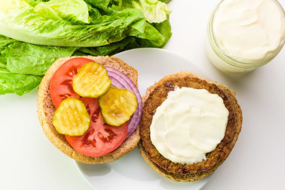 Looking down on a veggie burger with vegan mayonnaise. The other side of the bun has chopped tomatoes, onions, and pickles. There is a jar with more mayo and lettuce nearby.