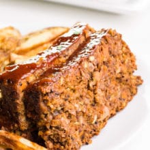 Two slices of vegan meatloaf sit beside French fries on a plate. Another plate of meatloaf is in the background.