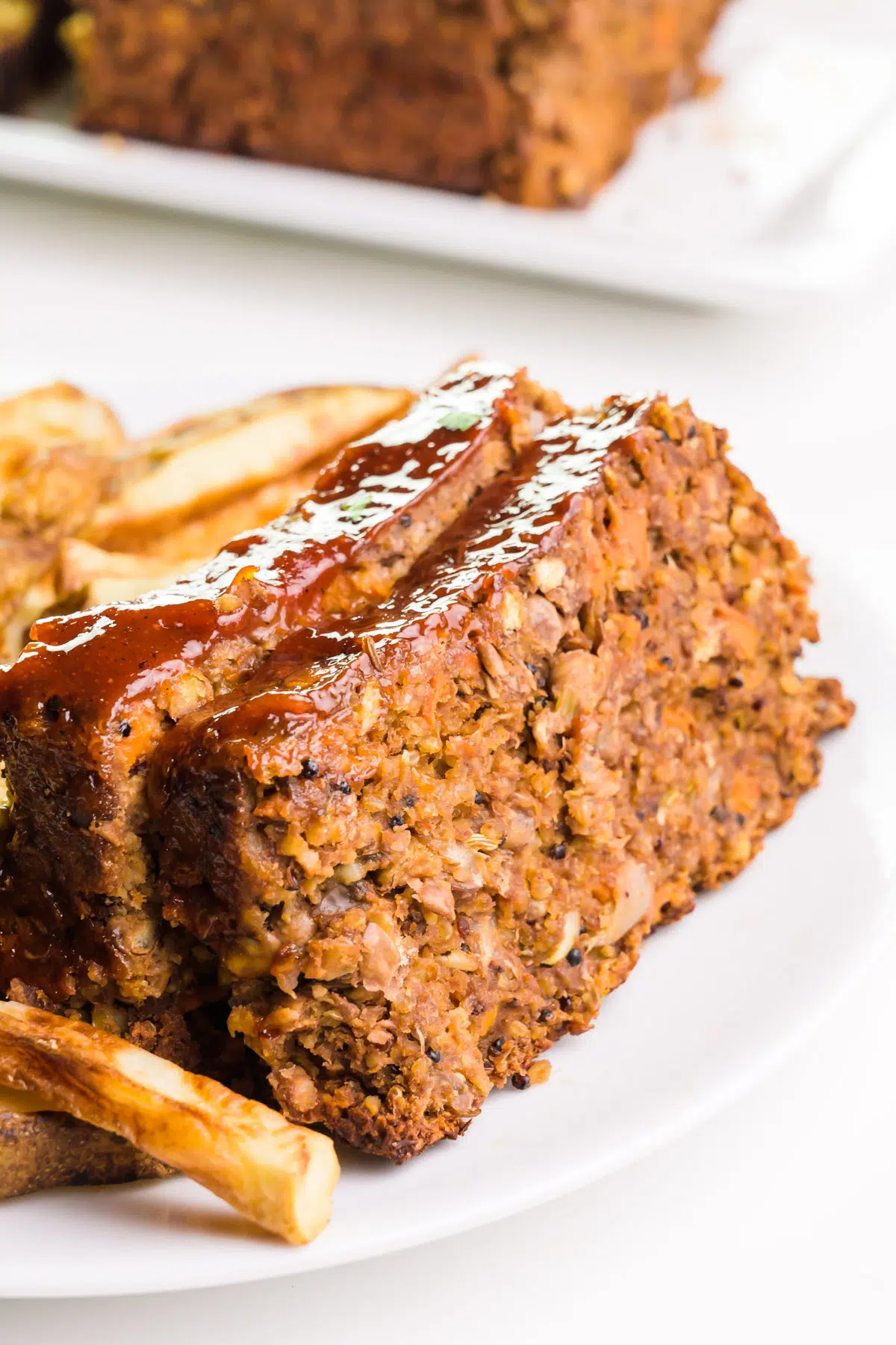Two slices of vegan meatloaf sit beside French fries on a plate. Another plate of meatloaf is in the background.