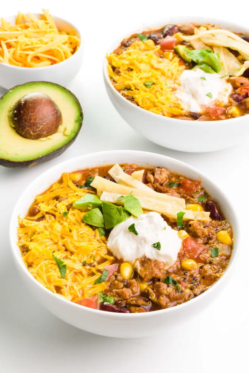 Two bowls of plant-based taco soup are loaded with toppings. There's a cut avocado and a bowl of vegan cheese in the background.