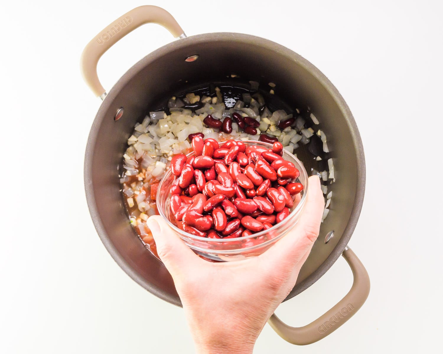 A hand holds a bowl of red kidney beans, pouring them into a pot with other ingredients.