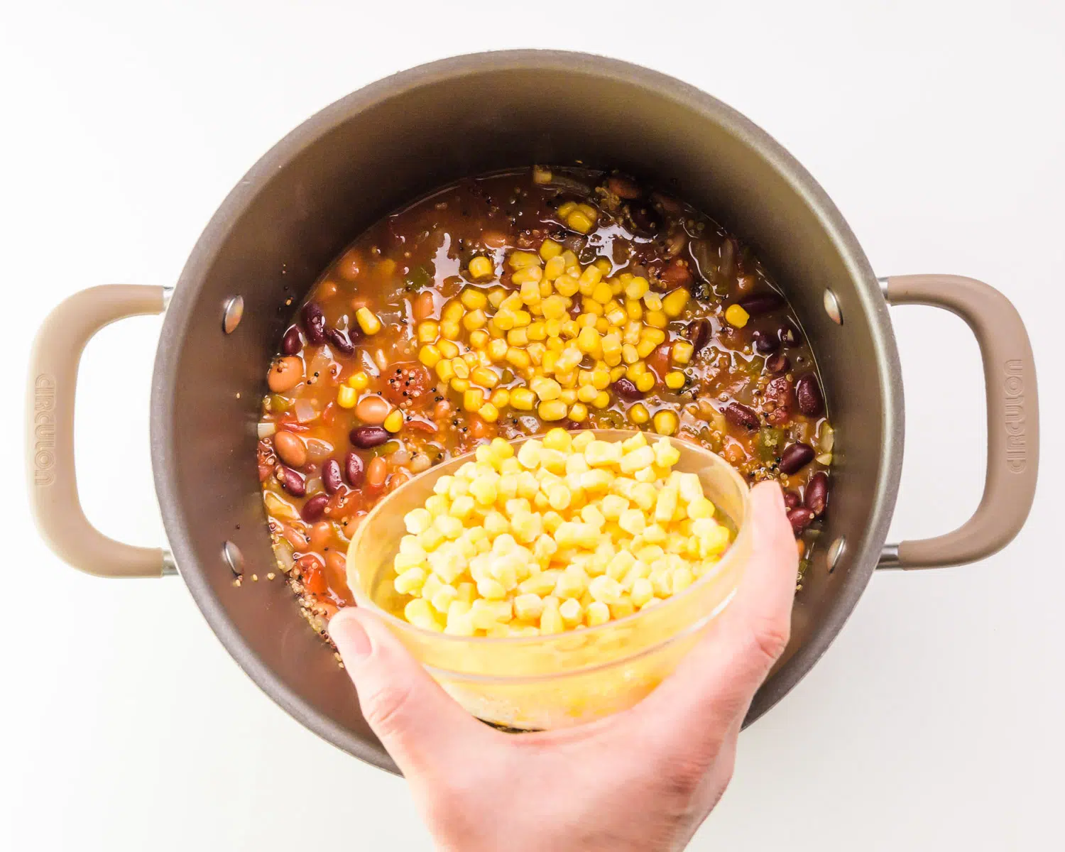 A hand holds a bowl of corn, pouring it into a pot with other soup ingredients.