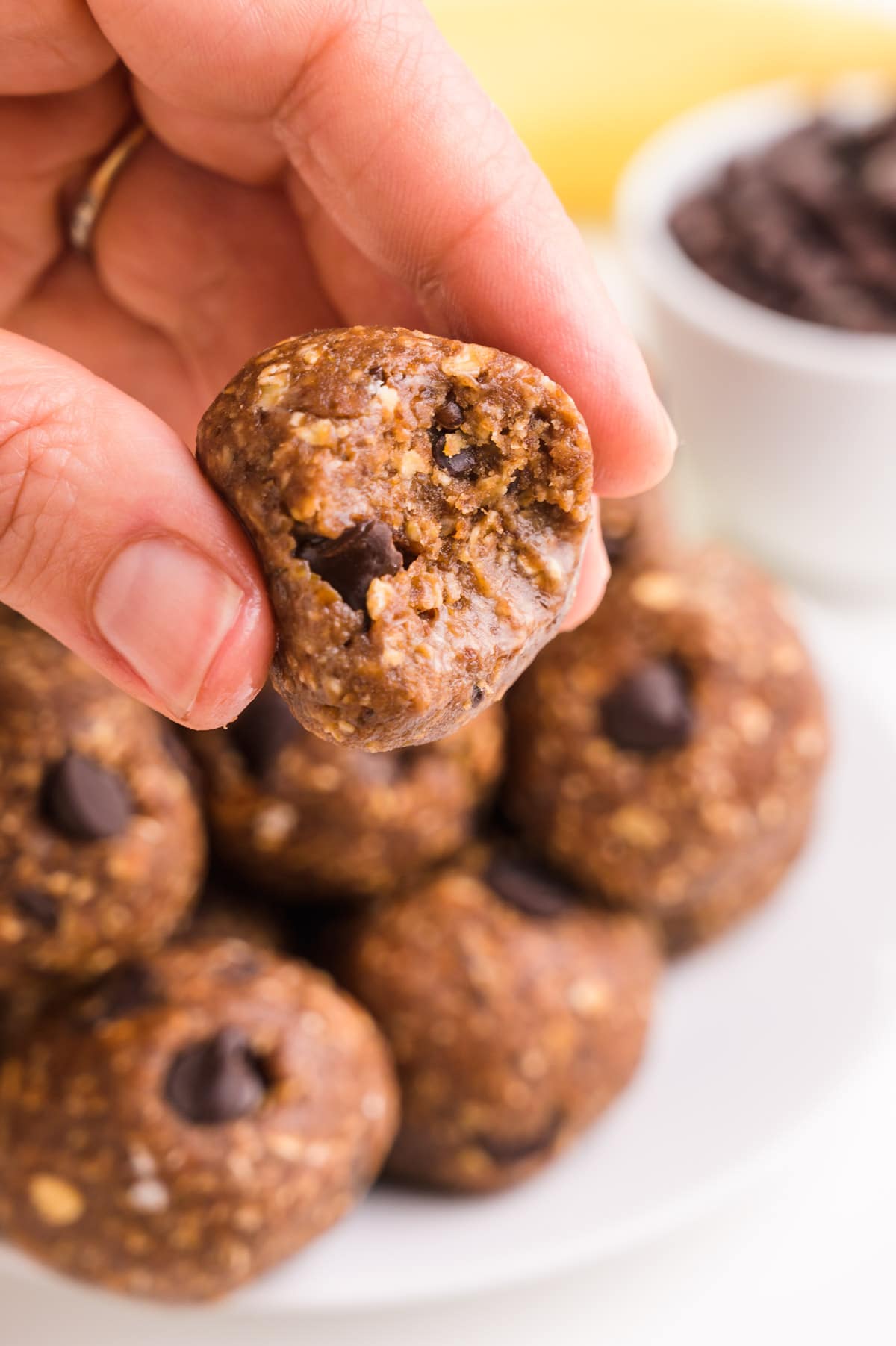 A hand holds a banana protein ball with a bite taken out. There are more in the background, along with chocolate chips and bananas.