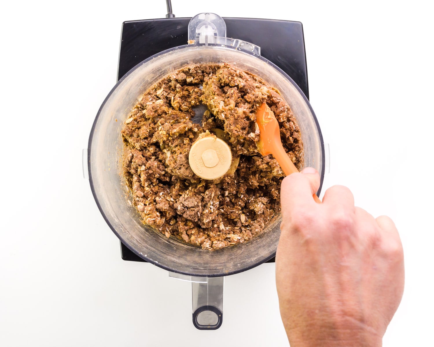 A hand holds a spatula, using it to push down ingredients in a food processor bowl.
