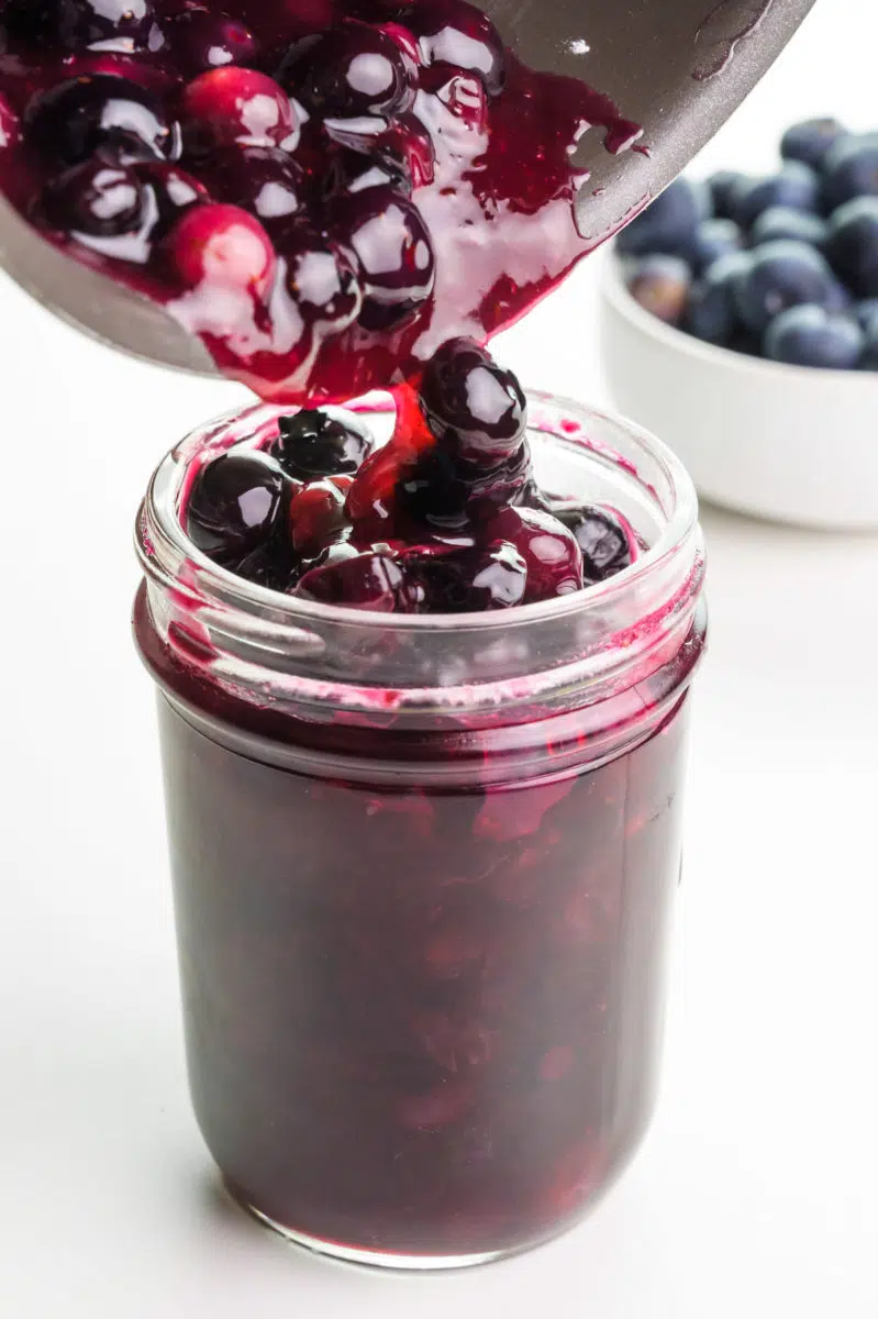 Blueberry glaze is being poured from a saucepan into a glass jar. There are fresh blueberries in the background.