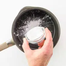 A hand holds a bowl of cornstarch, adding it to a saucepan.