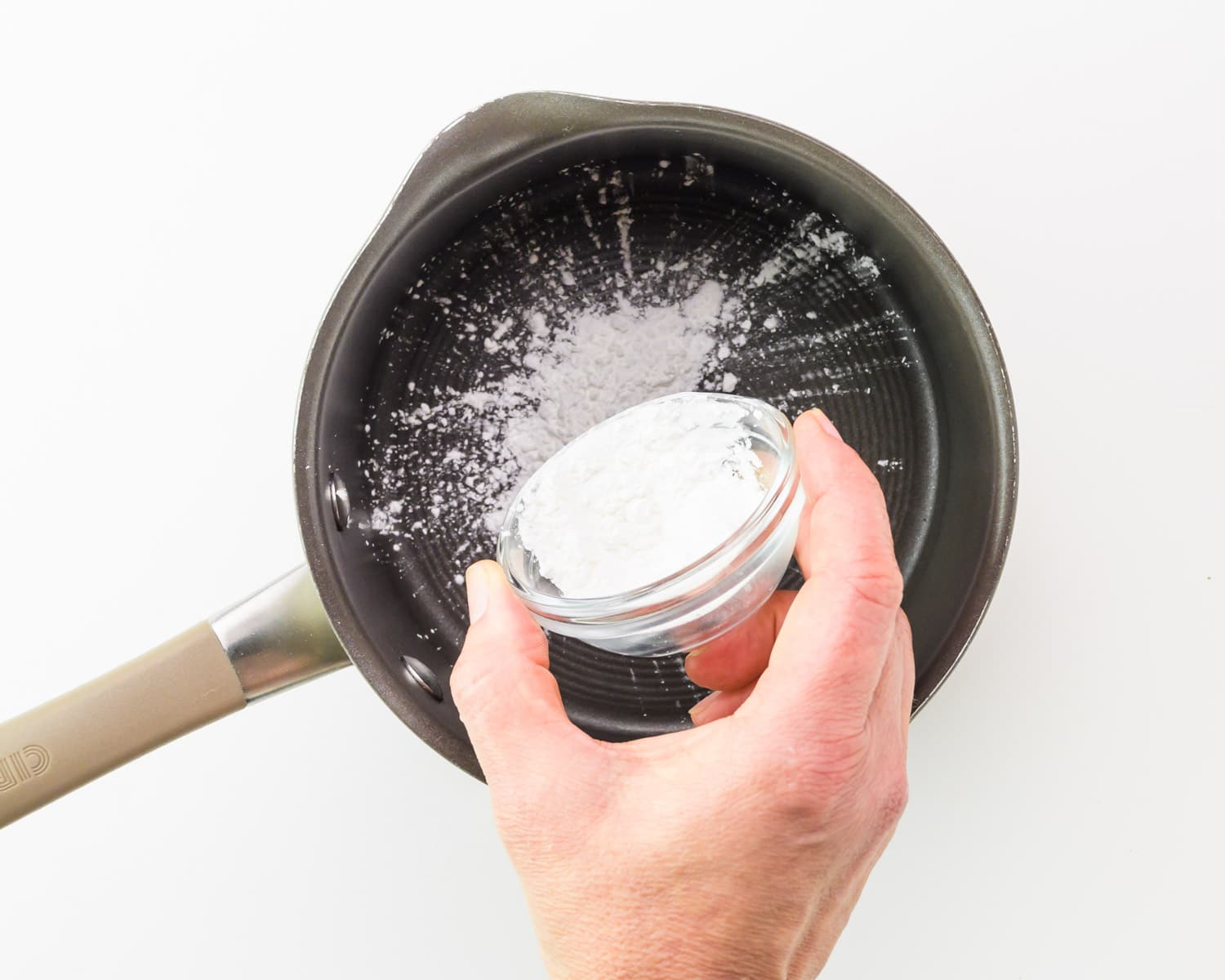 A hand holds a bowl of cornstarch, adding it to a saucepan.