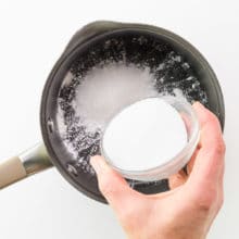 A hand holds a bowl of sugar, adding it to a saucepan with cornstarch.