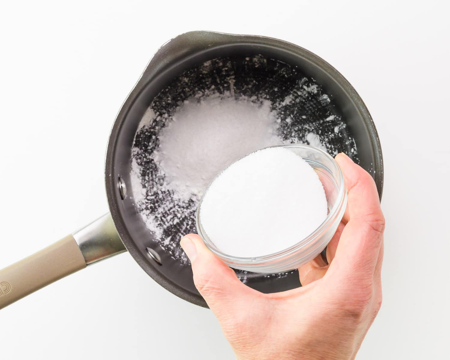 A hand holds a bowl of sugar, adding it to a saucepan with cornstarch.