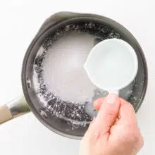 A hand holds a measuring cup of water, adding it to a saucepan with sugar and cornstarch.