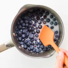A hand holds an orange spatula, using it to stir blueberries in a saucepan.
