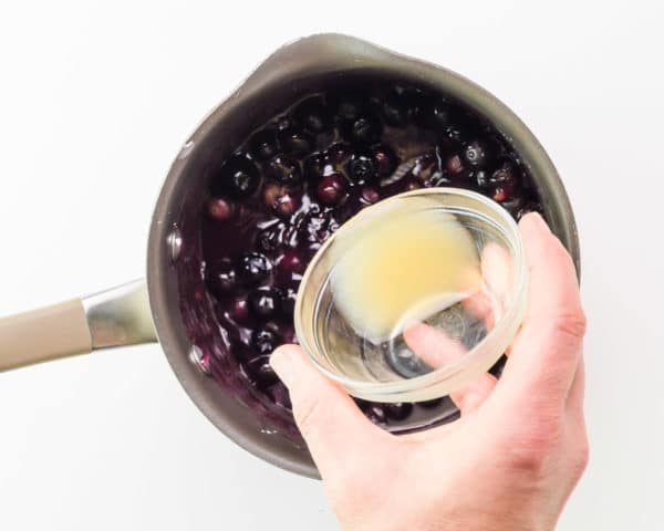 A hand holds a bowl of lemon juice, adding it to blueberry sauce in a saucepan.
