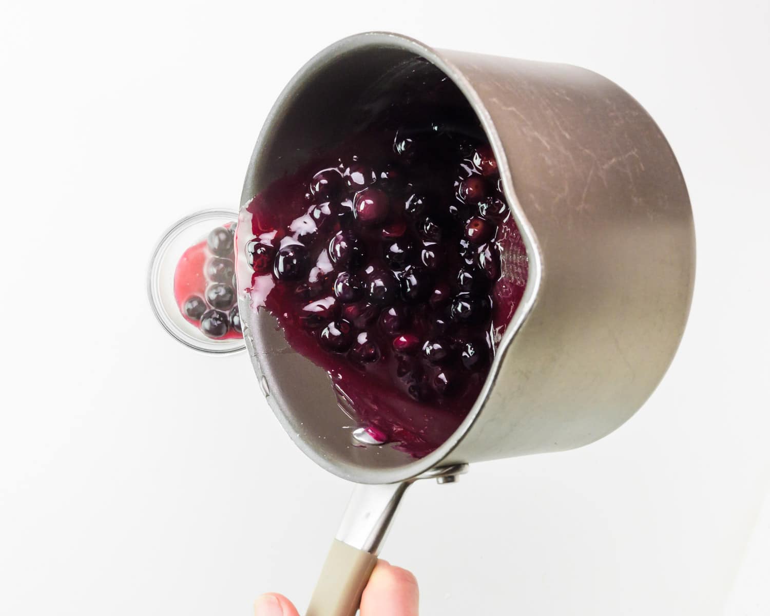 A hand holds a saucepan, pouring blueberry reduction into a glass jar.
