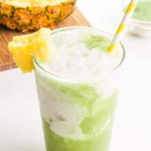A glass of pineapple matcha has a yellow straw in it. There is a bowl of matcha powder and a sliced pineapple on a cutting board in the background.
