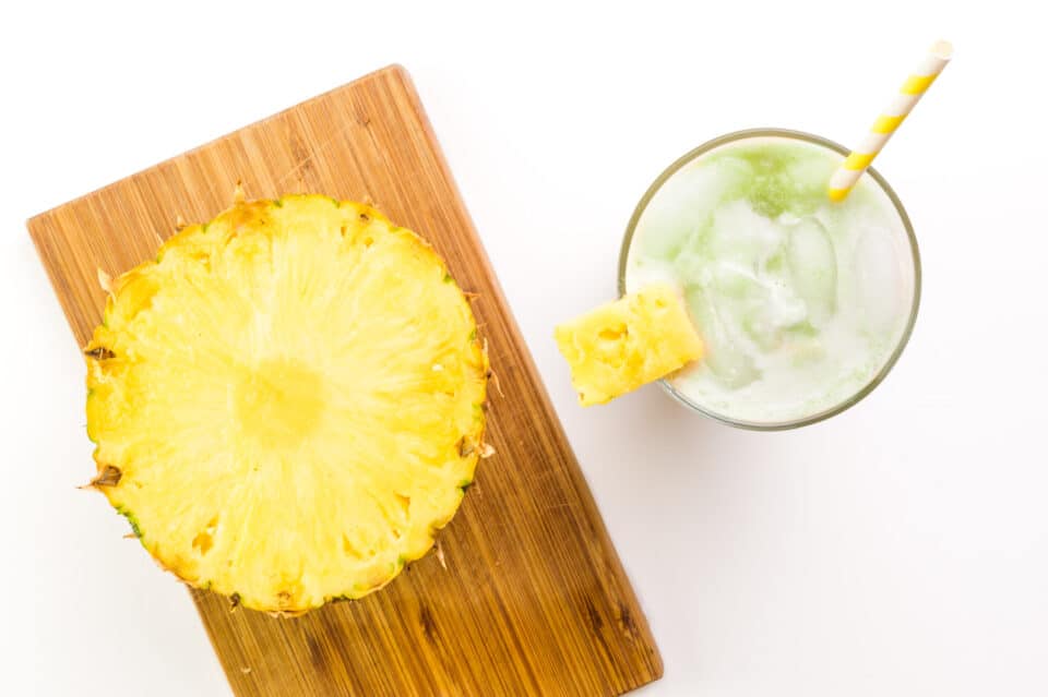 Looking down at a cut pineapple on a cutting board.  It sits next to a glass with an iced matcha drink inside.
